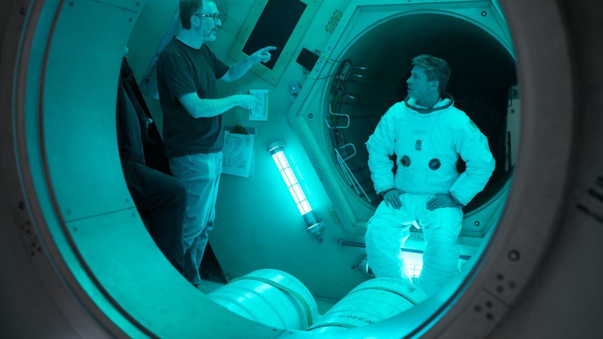 El director de la película James Gray, aparece a la izquierda de la imagen, viste pantalones vaqueros y una camiseta de color oscuro. Da indicaciones a Brad Pitt, que le escucha atentamente con los brazos en jarra sentado a poca distancia del director. Están en una sala preparada para el rodaje de una escena en una nave espacial. 